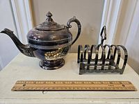 Silverplated teapot and toast caddy side by side with a ruler in front for scale.