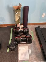 Two Bowflex SelectTech BD552 adjustable dumbbells on black trays with owner’s manual and boxed Pilates mat behind