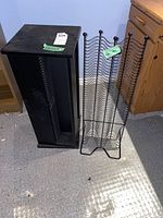 Two CD racks side by side on floor including a black wooden pressboard rack and a black wire rack with wavy design.