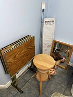 Lot corner showing retro folding TV trays on metal rack with wood grain laminate tops, adjustable wood stool, shower curtain rod in box, shoe rack boxed, small medicine cabinet mirror on floor