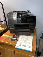 Full front view of HP Officejet Pro 8630 printer showing paper tray, display panel, power cord on top, extra ink cartridges, and manual sheets placed in front of printer.