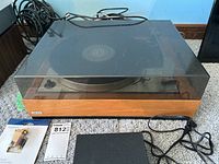 Overall front view of the Lenco B55 turntable with wood base, dust cover, and visible cables behind it. Insignia cartridge package and MaxSold lot tag placed beside the turntable.