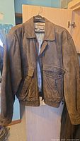 Brown Wilsons Leather jacket hanging on door, showing front with classic collar and two large front pockets.