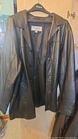 Front view of men's black leather jacket hanging on hanger, showing length, collar, and button closure