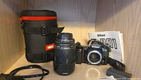 Nikon F-801 camera body without battery cover next to Tamron 70-300mm AF lens, carrying case and camera manual on shelf