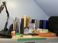 Overview of office items on shelf including clock, binders, task lamp base, post-it notes, file folders, Bluetooth earpiece and laptop bag