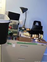 Photo of a black metal filing rack filled with rolls of tape next to a black desk lamp, stapler on top of file cabinet, and a waste bin at side.