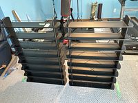 Pair of black shelving units standing side by side on carpet. Each unit has multiple shelves and wire crossbars on back. One wire crossbar is unattached but visible.