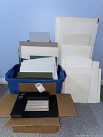Full view of the assortment of foam core boards and mattes in a blue rubbermaid bin and stacked beside it, various sizes and colors including white, grey, brown and green