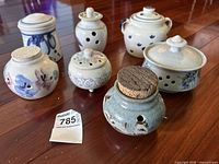 Set of seven signed pottery garlic bowls and small containers, arranged on wooden floor, showing decorative blue floral and ventilated designs. Two bowls have cork stoppers.