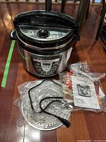 Full view of the pressure cooker with lid, power cord, manuals, and steaming rack visible on floor.