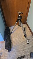 Full view of Manfrotto tripod set up with detachable extension leg and black soft carrying case placed beside it on carpeted floor.