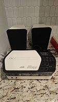 JVC stereo system unit with two speakers arranged behind the unit on granite countertop, showing top and front panels.