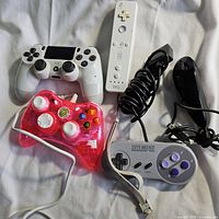 Photo showing all four controllers: a white Sony DualShock 4 wireless controller, a white Nintendo Wii Remote, a red translucent wired Xbox 360 controller, and a grey Super Nintendo controller. All controllers are placed on a white surface.