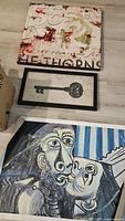 Three wall art pieces including a beige abstract painting with text, a framed black vintage key print, and a Picasso-style painting on canvas featuring two faces.