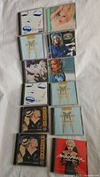 Photo showing ten Madonna CDs laid out on white fabric surface. Various album covers visible including multiple copies of the Fame album and others.