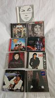 Photo shows 10 Michael Jackson music CDs in jewel cases laid out on white fabric background. Covers include various album artworks and styles.