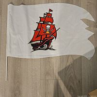 Tampa Bay Buccaneers flag with red pirate ship and skull logo on a white background, laid flat on a wooden floor.