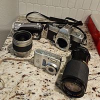 Lot showing four cameras and lenses on countertop, including Minolta SLR camera body, Kodak camcorder, Canon digital camera, and zoom lenses.