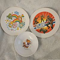 Two large melamine plates and one smaller bowl laid on fabric background showing Care Bears, G.I. Joe and a girl with monkey cartoon designs