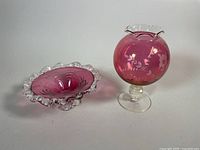 Photo showing two cranberry glass items: a bowl with scalloped rim and a pedestal globe vase with floral etching.