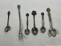 Six pieces of sterling silver cutlery laid on a white fabric background, showing spoons with decorative colored emblems on handles and a 2-prong fork