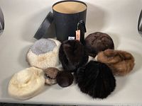 Photo showing six vintage fur hats, two fur ear-muffs and black cylindrical hat box.