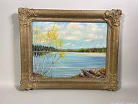 Front view of framed oil painting showing a lakeside scene with yellow-leaved birch trees, water, rocks and distant forest under a partly cloudy sky.