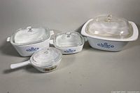 Four Corningware casserole dishes with clear glass lids arranged on a neutral surface, showing size and patterns.