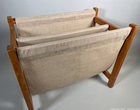 Front and side angled view of the teak and linen magazine rack showing fabric slings and wooden frame with visible wood grain and finish.