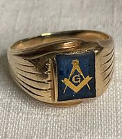 Front view of ring showing square bezel with blue enamel and gold Masonic square and compass emblem, with engraved shoulders.