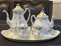 Five-piece porcelain tea set on black surface, includes coffee pot, teapot, sugar bowl, creamer, and oval serving tray with gold hand painted designs