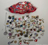 Wide shot showing a red hat covered in assorted pins with many additional pins laid out beside it in rows on a white surface along with a silver necklace with an engraved pendant.