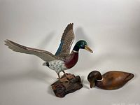 Front and side view of two wooden mallard duck sculptures: one flying pose on wood base, one simple carved duck without base