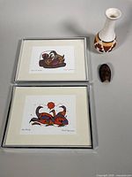 Photo of two framed Norval Morrisseau prints, signed, 9x12 inches each, showing vibrant Indigenous art images with earth tones and black outlines.
