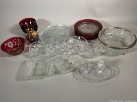 Wide view showing the entire collection of red ruby and clear crystal dishes on a white surface including serving bowls, individual bowls, plates, trays, and small dishes