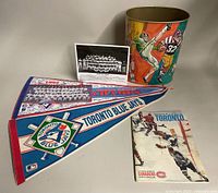 Metal trash can with colorful sports player graphics alongside two Toronto Blue Jays pennants, 1982 New York Yankees team photo, and another pennant.