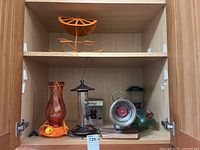 Cabinet shelf showing orange metal garden décor and three hummingbird feeders including clear glass and orange plastic models, vintage Powell & Hanmer bicycle lamp on wooden base, Coleman lantern and boxed fondue set.