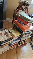 Stack of assorted hardcover and softcover books piled on the floor beside a wooden stool with a woven seat.