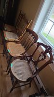 Four wooden antique dining chairs with caned seats lined up by a window, showing some wear on the wood finish and woven cane seats.