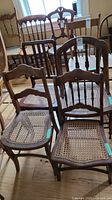 Photo showing four wooden dining chairs with caned seats arranged on a wood floor. Chairs have varying carved back designs and show signs of wear on the wood finish. Seat cane is intact on all chairs.