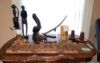 Wide view of the collection of wooden carvings including figurines, carved panel, candle holders, and letter opener, arranged on a polished wooden table near window.