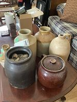 Full view of six stoneware crocks and jugs including black crock with chip and brown bean pot with lid on wooden table in living room.
