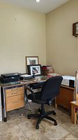 Desk with three drawers on the left, glass top cover, various items on top including printer and framed pictures; black swivel office chair positioned in front