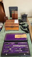 Vintage Hughes-Owens Co. drafting set open showing tools in black case with purple lining, wooden box, watches, and other wooden items on green table