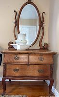 Front view of vintage wooden dresser with three drawers and attached oval wooden mirror. Decorative metal drawer pulls and carved wood details are visible. White ceramic pitcher and basin are placed on top of the dresser.