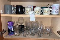 Shelf with 16 coffee mugs on top and assorted clear water glasses on bottom including tumblers and short glasses.