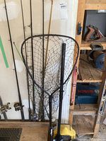 View showing four fishing rods with reels leaning against wall with fishing net, paddle, minnow pail and rubber boots visible nearby.
