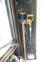 Photo showing pitch fork with blue handle grip and a shovel with yellow grip placed upright inside a shed