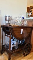 Wooden serving cart with glass and stainless steel items on and under it including stemmed glasses, mugs, salad bowls, fondue pot, teapots, wine decanter and a stainless pot.